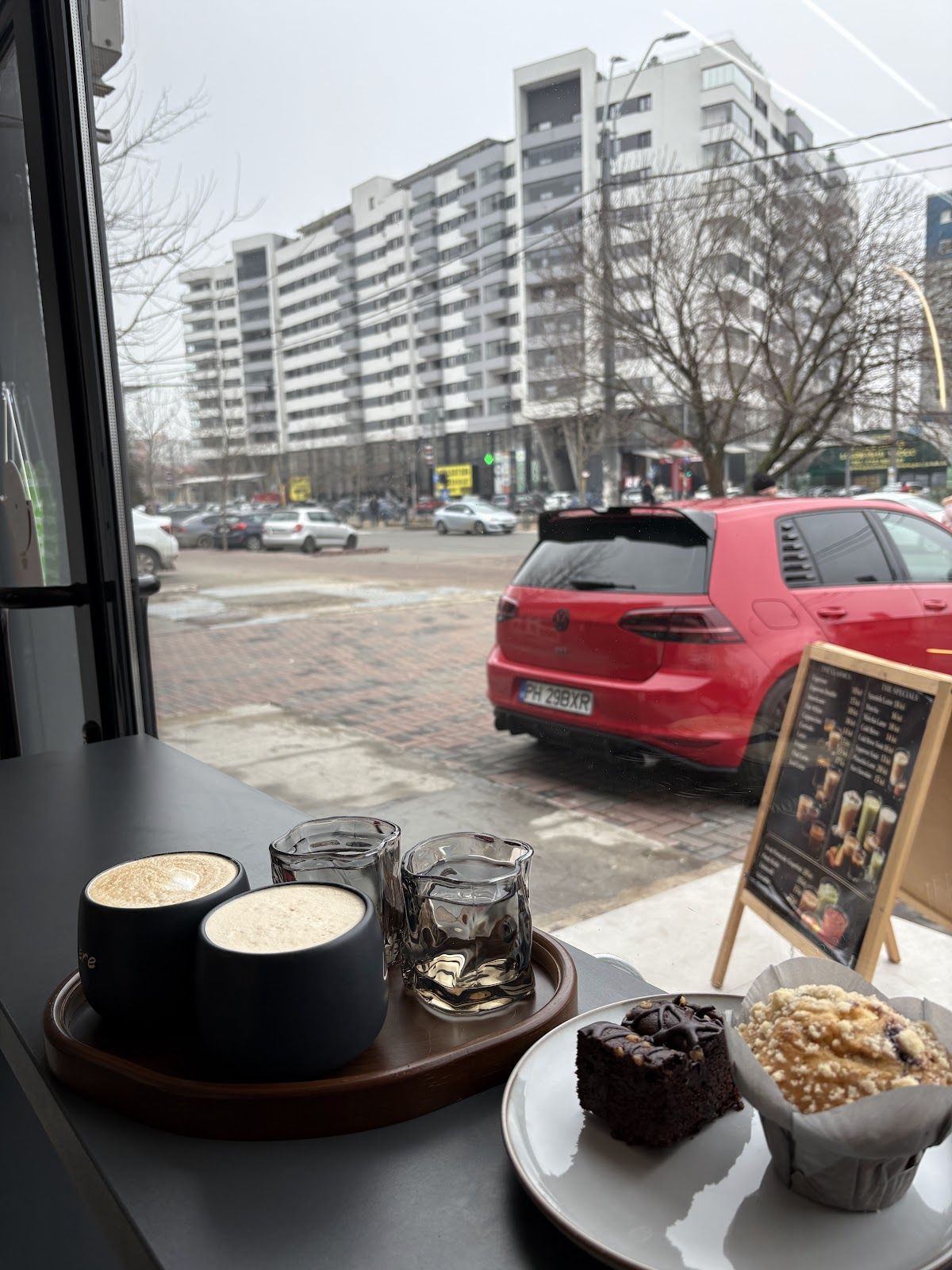 Latte pe tavă de lemn cu brownie și muffin la fereastra Lumière, cu priveliștea spre Șoseaua Mihai Bravu